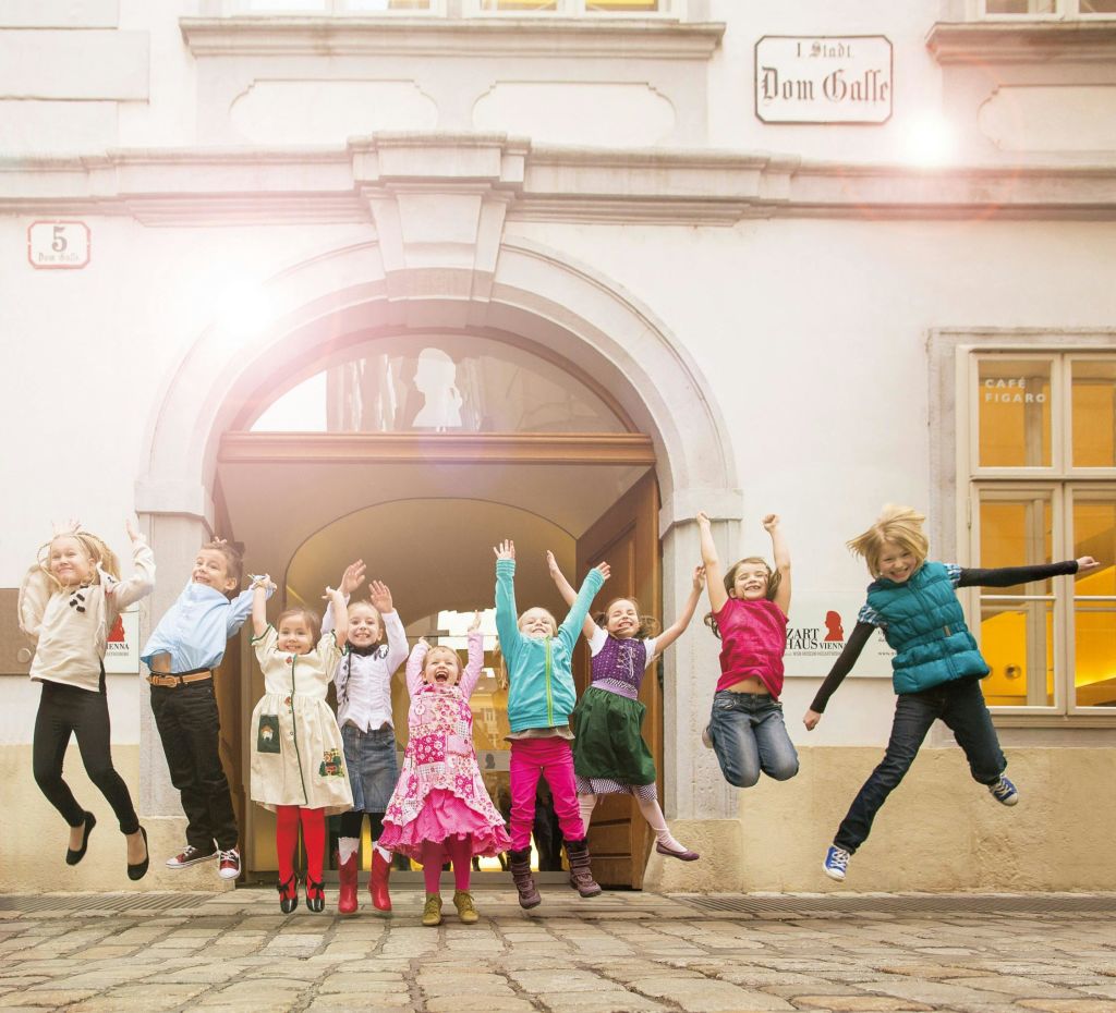9명의 어린이가 자갈 마당의 아치형 출입구 앞에서 즐겁게 뛰어놀고 있으며, 위에는 "Dom Galle" 이라고 적힌 표지판이 있습니다.