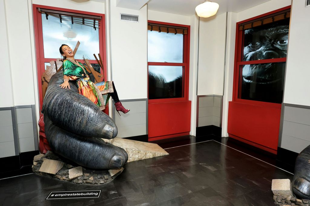 빨간색 프레임의 창문이 있는 방에는 화려한 색상의 옷을 입은 사람이 고릴라의 손을 닮은 커다란 소품에 앉아 있습니다.