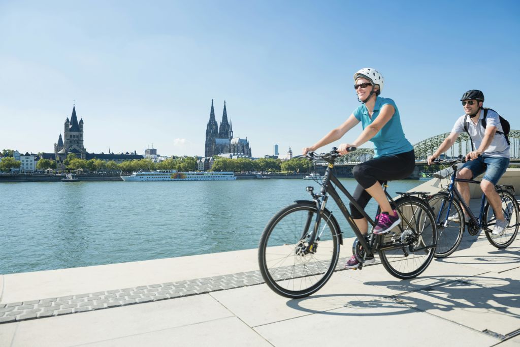 두 사람이 강변 길을 따라 자전거를 타고 있습니다. 맑고 푸른 하늘 아래 배경으로 대성당과 다리가 보입니다.