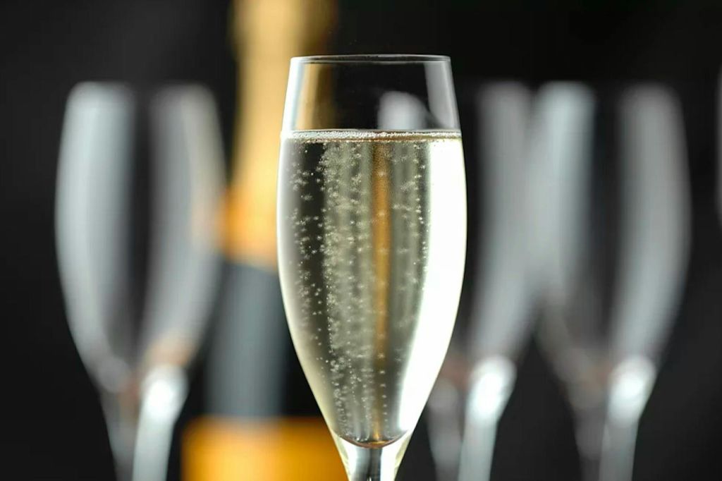 흐릿한 샴페인 병과 잔을 배경으로 스파클링 와인이 가득한 샴페인 플루트를 클로즈업한 사진입니다.