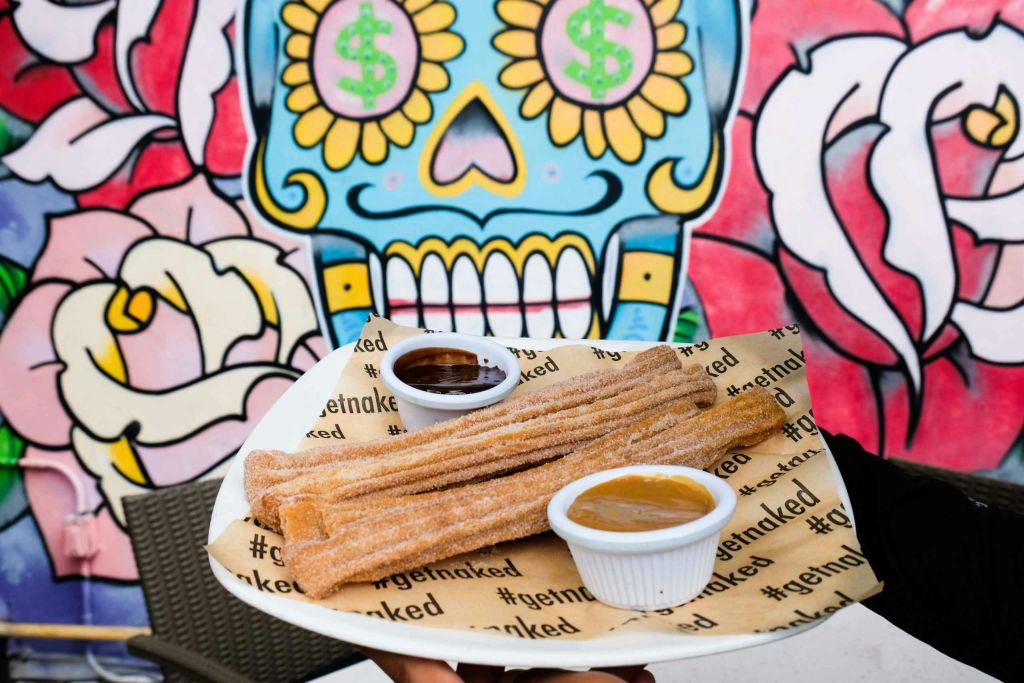 색색의 벽화 앞에 담그는 소스를 곁들인 츄러스 한 접시. 파란색 두개골에 머니 사인이 그려져 있습니다.