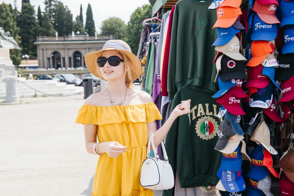 화창한 날 야외 시장에서 노란색 오프숄더 드레스와 밀짚 모자를 입은 여성이 쇼핑을 하고 있습니다.