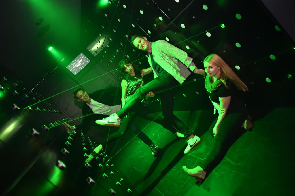 장난스럽고 일그러진 시각으로 녹색 악센트 조명이 있는 거울 벽을 배경으로 활기차게 포즈를 취하는 세 사람.