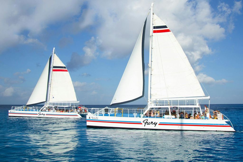 붉은색, 검은색, 파란색 줄무늬의 흰색 쌍동선 두 마리가 부분적으로 흐린 하늘 아래 잔잔한 바다를 항해합니다. 직원이 온보드에 표시됩니다.