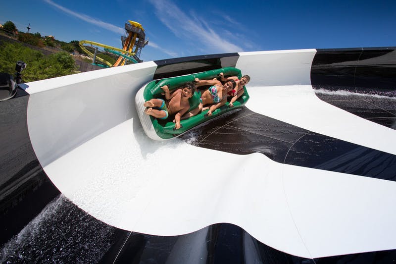 푸른 하늘 아래 흰색과 검은색의 커다란 워터 슬라이드에서 녹색의 팽창식 뗏목을 타고 네 명이 함께 타고 있습니다.