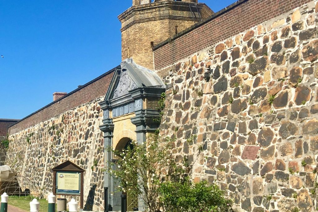 아치형 입구, 정보 표지판, 맑고 푸른 하늘 아래 녹지가 있는 유서 깊은 벽돌과 석조 건물 외관.