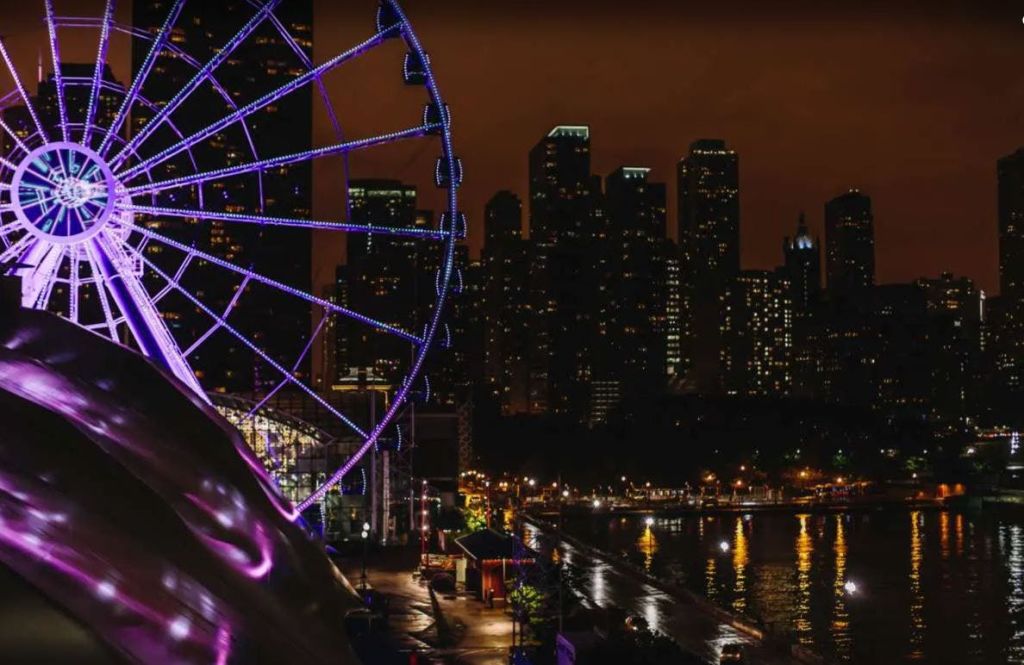 해안가 옆에 보라색 조명이 있는 대형 대관람차로 밤에는 도시의 고층 빌딩을 배경으로 조명이 켜져 있습니다.
