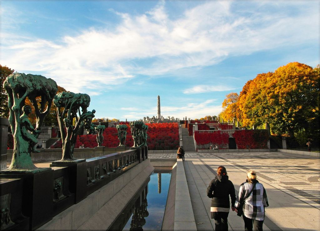 햇살이 내리쬐는 푸른 하늘 아래, 가을 단풍이 물씬 풍기는 조형물과 나무가 늘어선 통로를 따라 걷는 사람들.