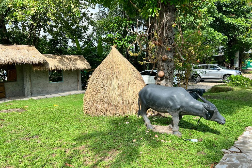 잔디가 우거진 지역에 있는 물소의 검은 조각상과 인접한 조롱박이 걸려 있는 나무 근처의 잔디 오두막과 초가 지붕 쉼터.