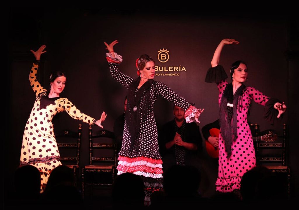무대에서 플라멩코를 연주하는 폴카 도트 드레스를 입은 두 명의 여성이 기타리스트와 클래퍼를 배경으로 합니다. 텍스트는 "Bulería" 입니다.