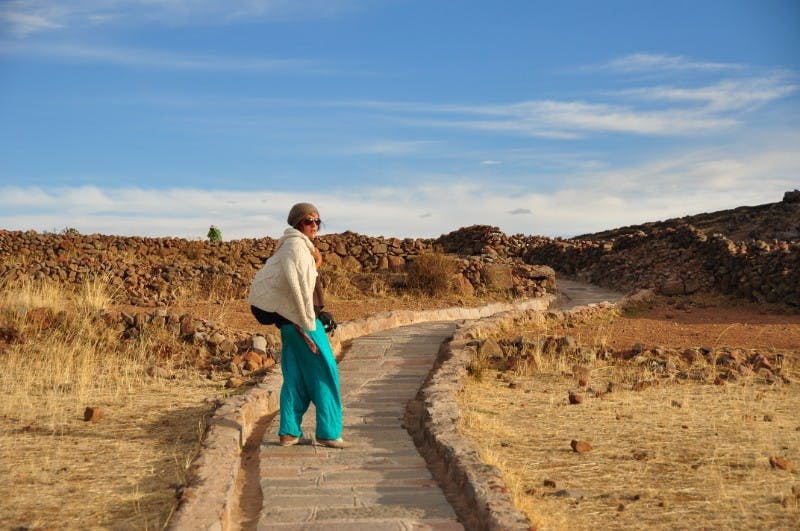 청록색 바지와 흰색 숄을 입은 사람이 맑고 푸른 하늘 아래 건조하고 바위가 많은 풍경의 돌길에 서 있습니다.