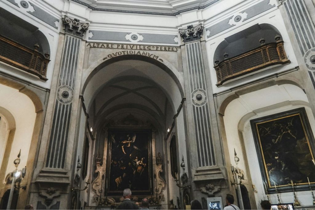 벽에 높은 아치, 기둥, 라틴 문구가 새겨진 화려한 예배당 내부에서 예술품을 감상하는 사람들.