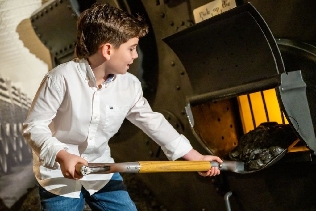 흰색 셔츠와 파란색 청바지를 입은 어린 소년이 긴 손잡이 도구를 사용하여 전시 또는 전시물과 상호 작용합니다.