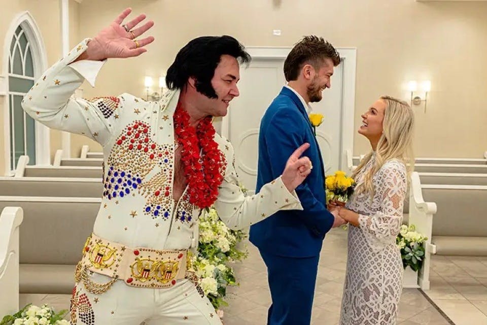 결혼식 복장을 한 커플이 예배당에서 서로 마주보고 서 있고, 엘비스 프레슬리 (Elvis Presley) 의 옷을 입은 한 남자가 근처에서 열정적인 포즈를 취하고 있습니다.