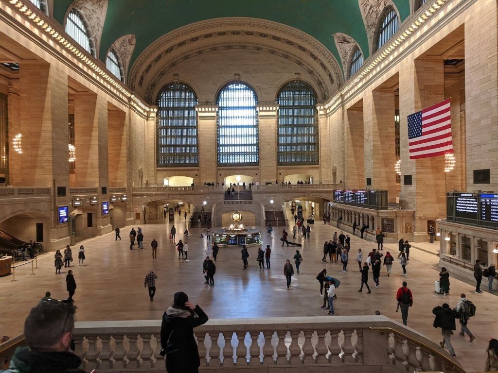 그랜드 센트럴 터미널의 메인 중앙 홀에는 높은 아치형 창문, 상징적인 시계, 걷는 사람들, 미국 국기가 있습니다.
