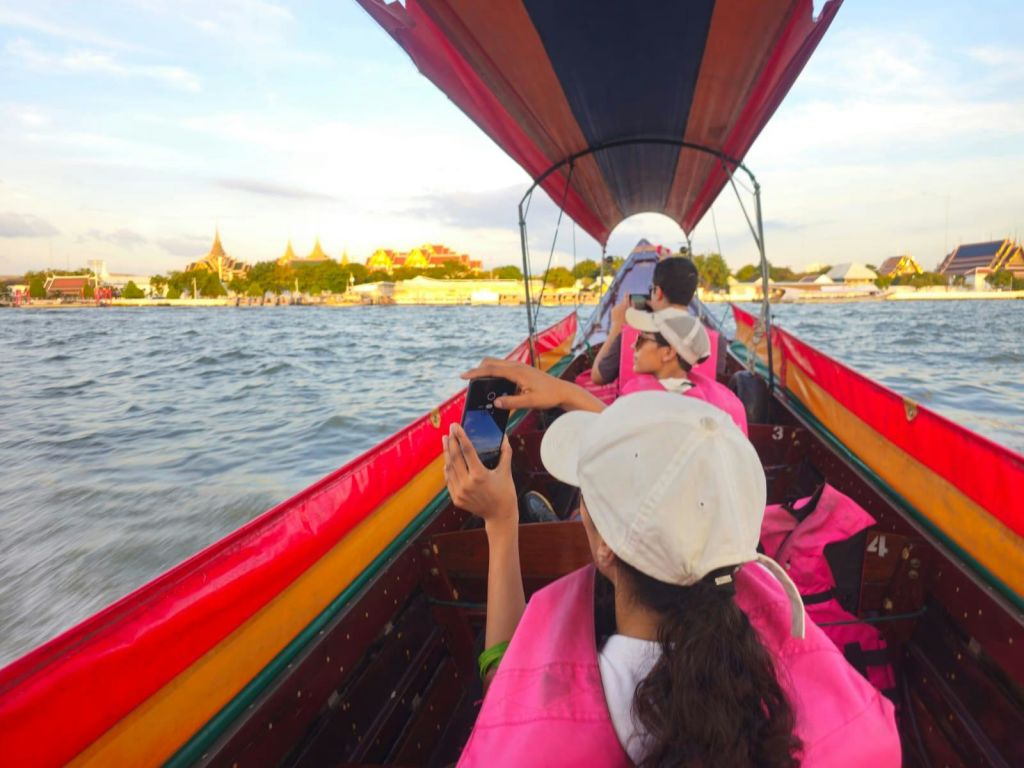 분홍색 구명조끼와 흰색 모자를 쓴 사람들이 강가에서 보트에 앉아 저 멀리 보이는 황금빛 사원 구조물 사진을 찍고 있습니다.