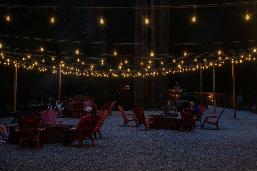 나무로 둘러싸인 자갈밭에서 밤에 줄 조명 아래 화덕 주위에 빨간 의자에 앉아 있는 사람들.