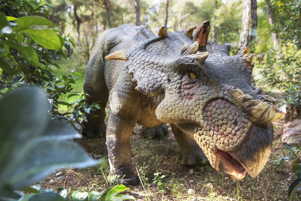 녹색 단풍으로 둘러싸인 숲 속에 질감이 살아있는 피부와 뿔이 달린 얼굴을 한 생생한 공룡 모델이 서 있습니다.