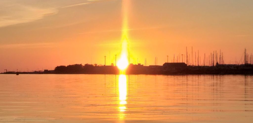 잔잔한 물 위에 해가 지고 황금빛으로 빛나며 건물과 기둥의 실루엣으로 표면을 비춥니다.