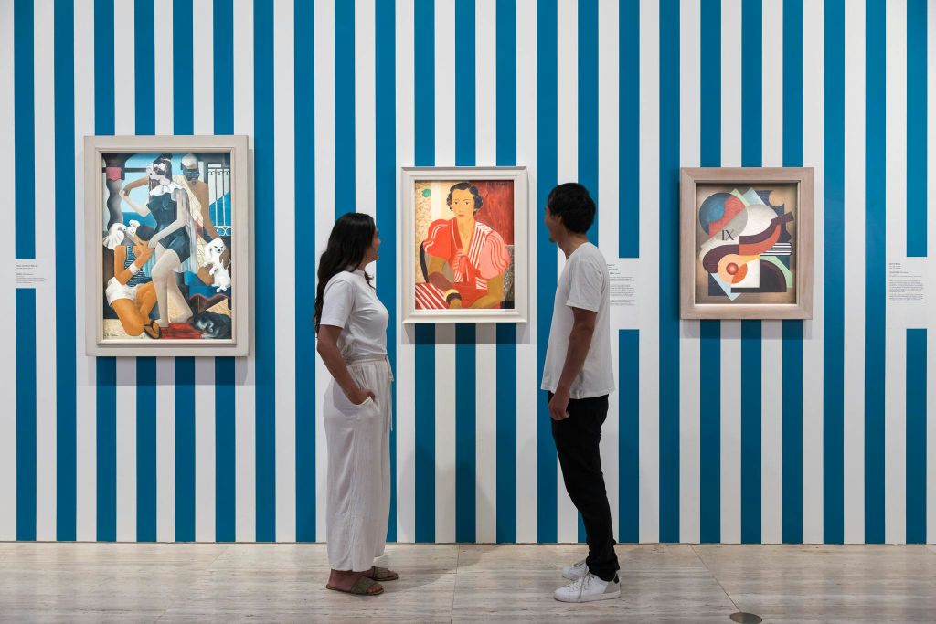 두 사람이 갤러리를 배경으로 파란색과 흰색 세로 줄무늬가 있는 벽에 걸린 그림을 관찰합니다.