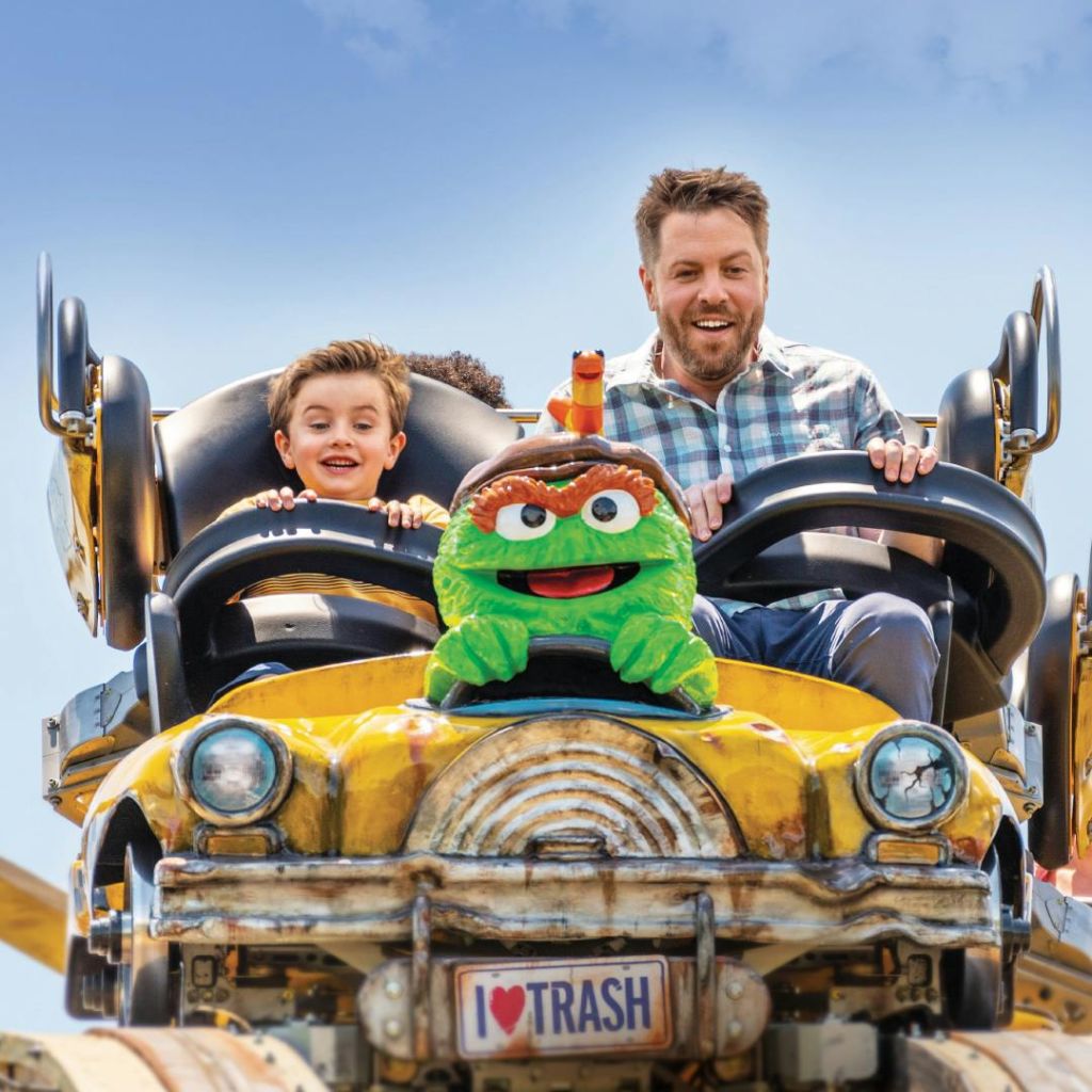 한 남자와 소년이 녹색 캐릭터와 "I ♥ Trash" 번호판을 테마로 한 노란색 롤러코스터를 신나게 타고 있습니다.