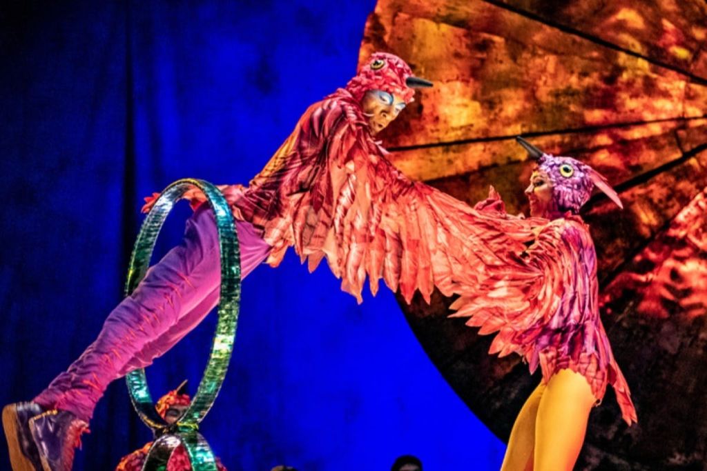선명한 색상의 새를 주제로 한 의상을 입은 두 명의 공연자가 무대 위의 원형 장치에서 곡예 연기를 펼칩니다.