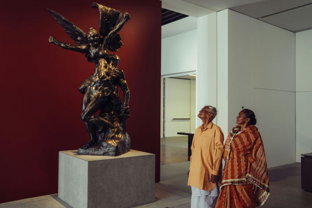 짙은 붉은 벽에 세워진 박물관에서 두 노인이 커다란 청동 동상을 감상하고 있습니다.
