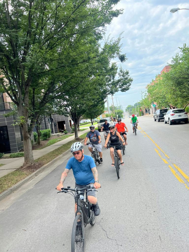 나무와 주차된 차량이 늘어선 교외 거리에 자전거를 타고 있는 한 무리의 사람들.