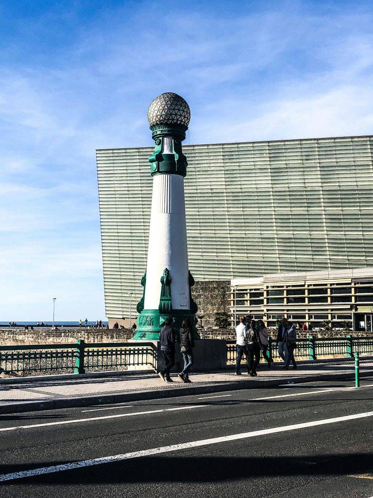 사람들은 커다란 현대식 건물과 바다를 배경으로 다리의 높고 화려한 녹색과 흰색 가로등을 지나갑니다.