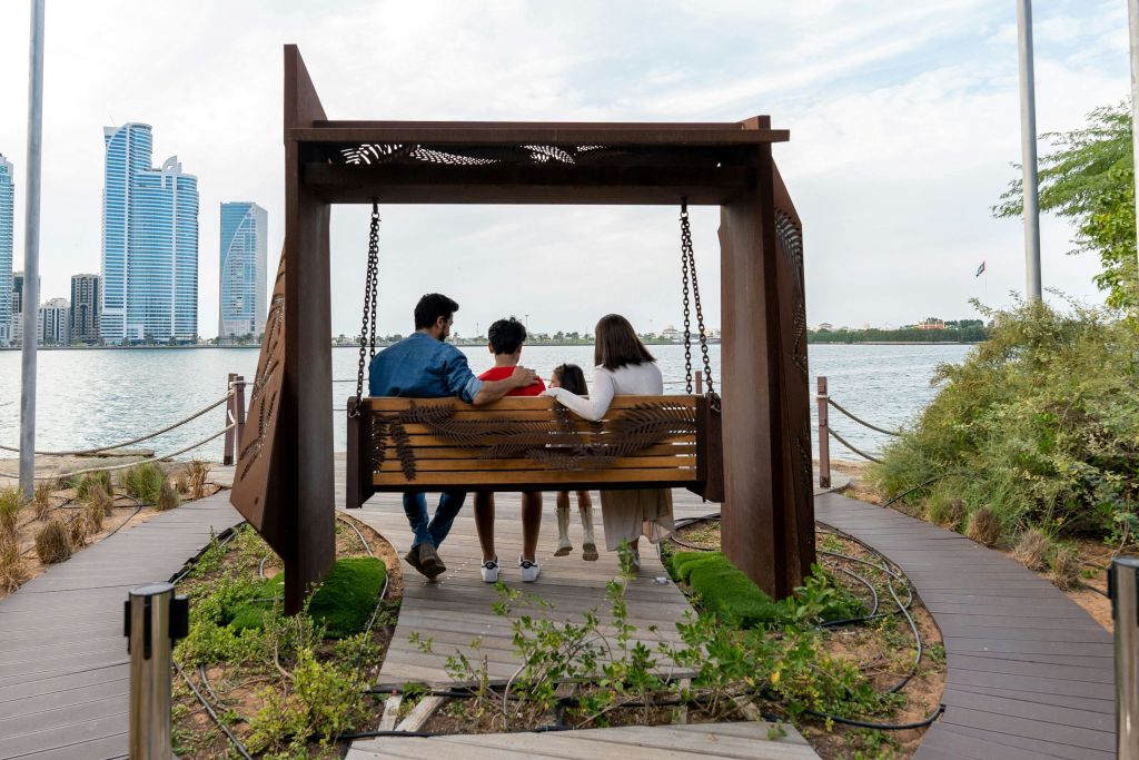 현대적인 건물과 녹지를 배경으로 세 사람이 나무 그네 벤치에 앉아 호수를 바라보고 있습니다.