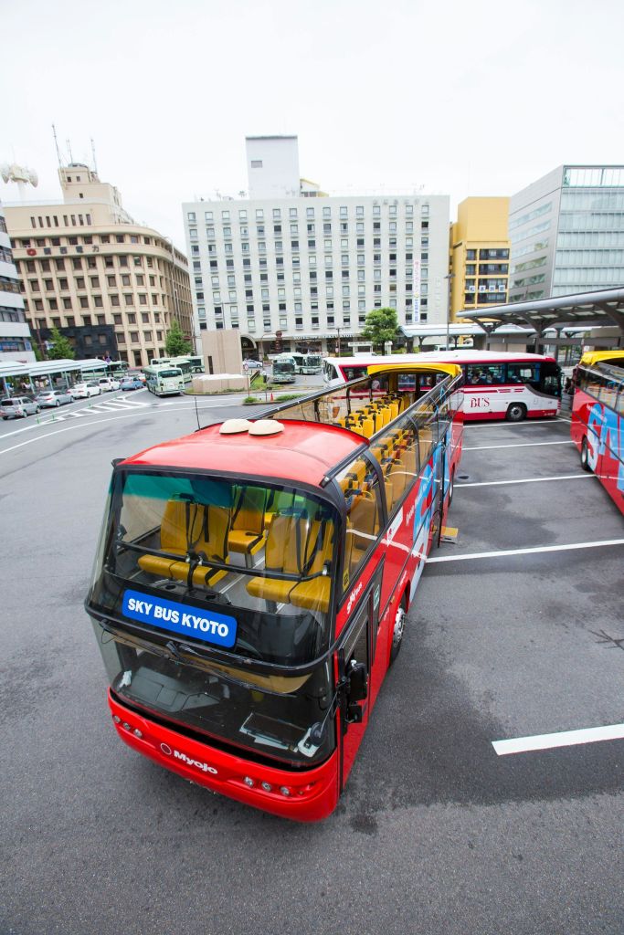 '스카이 버스 교토' 라는 이름의 빨간색 2층 버스가 비슷한 버스와 건물이 여러 개 있는 버스 주차장에 주차되어 있습니다.