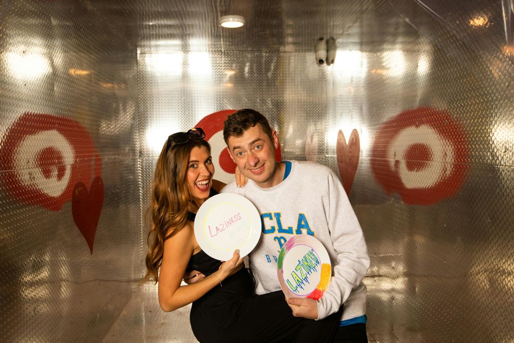 "게으름" 이 적힌 접시를 들고 미소를 짓고 있는 두 사람이 붉은 하트로 장식된 금속으로 된 방에 서 있습니다.