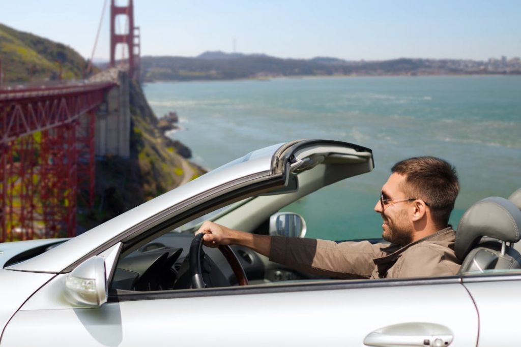 선글라스를 쓴 남성은 해변에서 컨버터블 차량을 운전하며 바다와 먼 언덕을 배경으로 아름다운 풍경을 감상합니다.