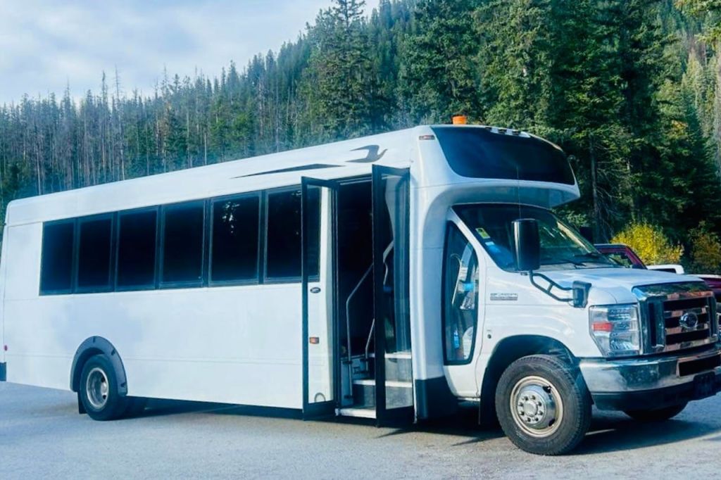 탁한 하늘 아래 빽빽한 상록수로 둘러싸인 도로에 열린 문이 있는 흰색 셔틀버스.