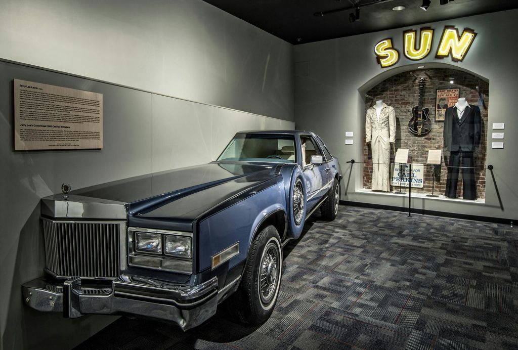 흰색 정장과 기타를 입은 마네킹 근처의 박물관에 파란색 클래식 자동차가 전시되어 있습니다. "태양" 네온 사인이 부분적으로 보입니다.