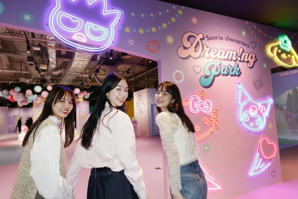 네온사인과 장난기 넘치는 장식이 돋보이는 다채로운 실내 전시장에서 세 명의 여성이 미소를 지으며 포즈를 취하고 있습니다.