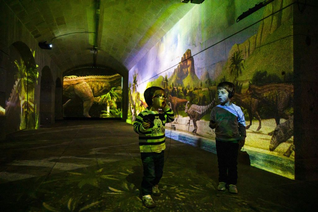 벽에 공룡이 투영된 조명이 희미한 방에 서 있는 두 어린이는 몰입감 넘치는 선사 시대 환경을 조성합니다.