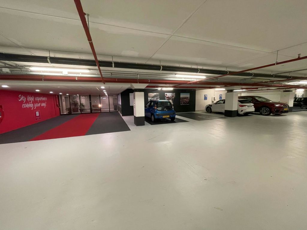 파란색 차량과 흰색 차량 2대가 주차된 주차장이 있습니다. 빨간색과 회색 카펫은 유리로 된 양문 입구로 이어집니다.