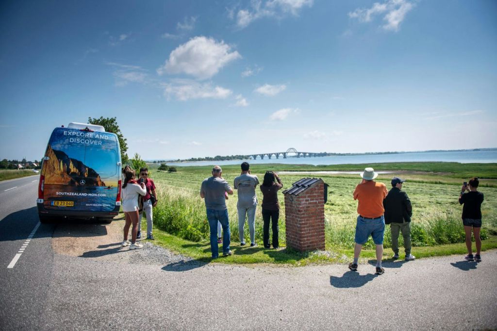 한 무리의 사람들이 주차된 밴 근처에 서서 맑은 하늘 아래 물 위로 다리가 놓인 경치 좋은 풍경을 감상하고 있습니다.