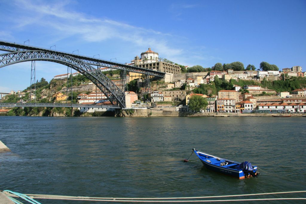 푸른 보트가 커다란 철교 근처의 강에 떠 있으며, 화려한 건물의 언덕과 교회를 배경으로 합니다.