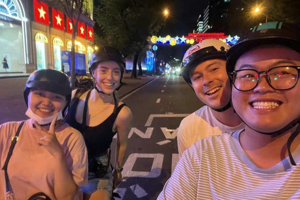 축제 장식과 도시 건물을 배경으로 밤에 불이 켜진 거리에서 헬멧을 쓰고 미소 짓는 세 사람이 포즈를 취하고 있습니다.