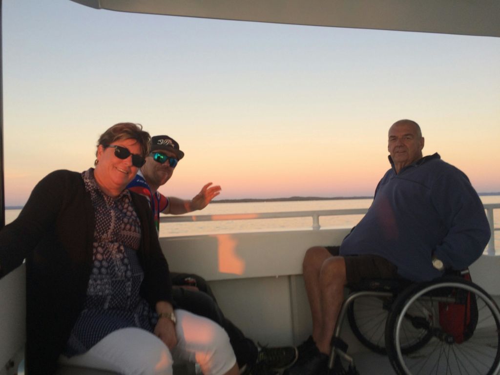 휠체어에 탄 사람을 포함해 3명이 해질녘 보트에 앉아 잔잔한 물놀이를 즐기고 있습니다.