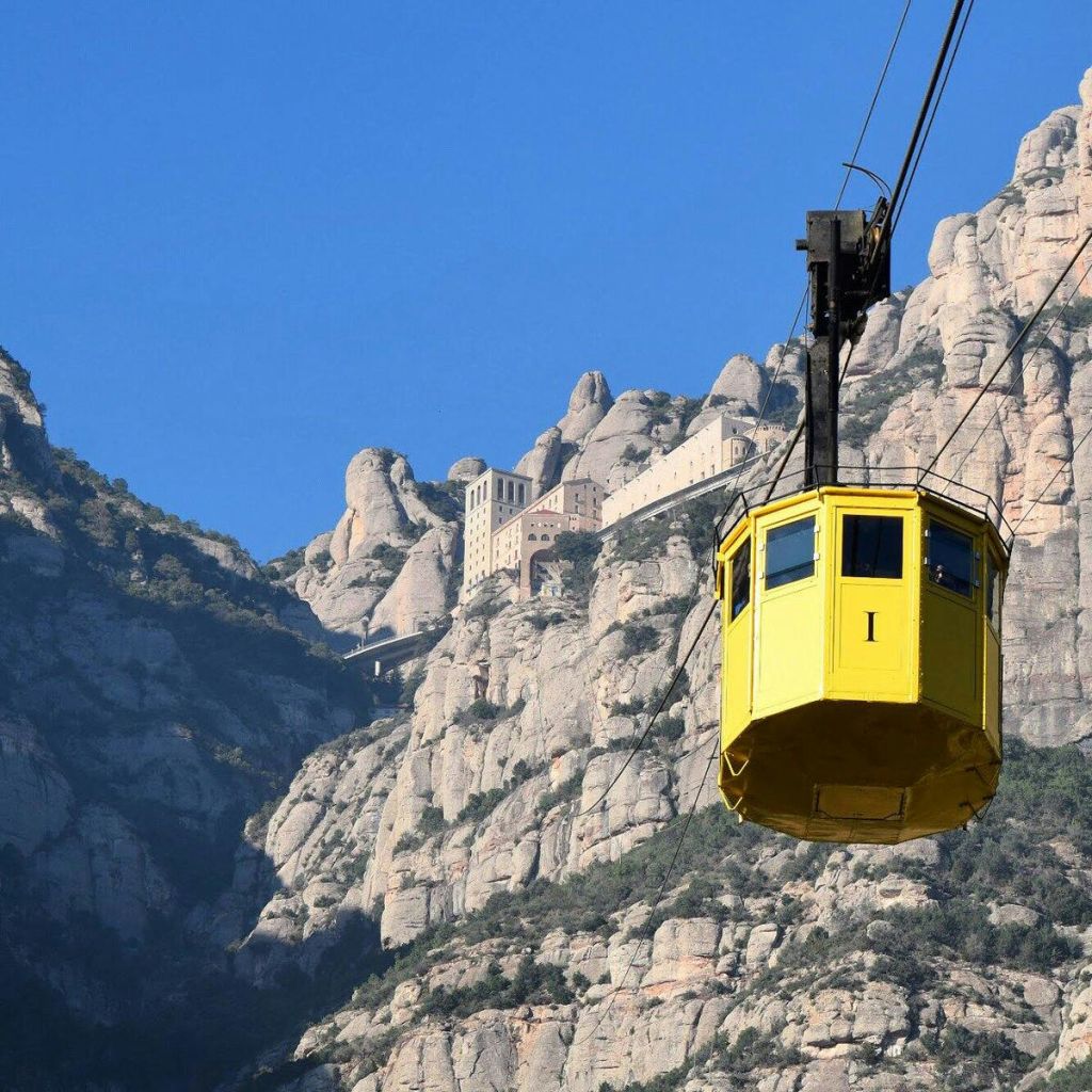 노란색 케이블카가 맑고 푸른 하늘 아래 험준하고 바위 같은 산들 사이에 자리 잡은 수도원을 향해 올라갑니다.