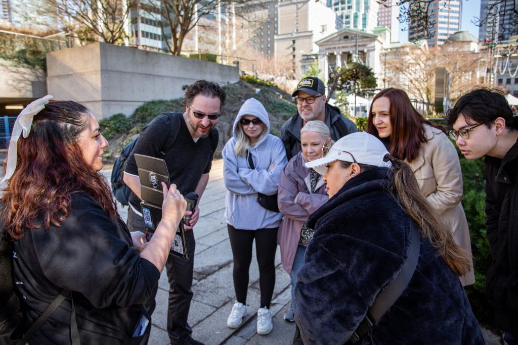 여섯 명의 사람들이 한 여성이 들고 있는 물건을 세심하게 바라보며 야외에 모였습니다. 도시 건물이 배경입니다.