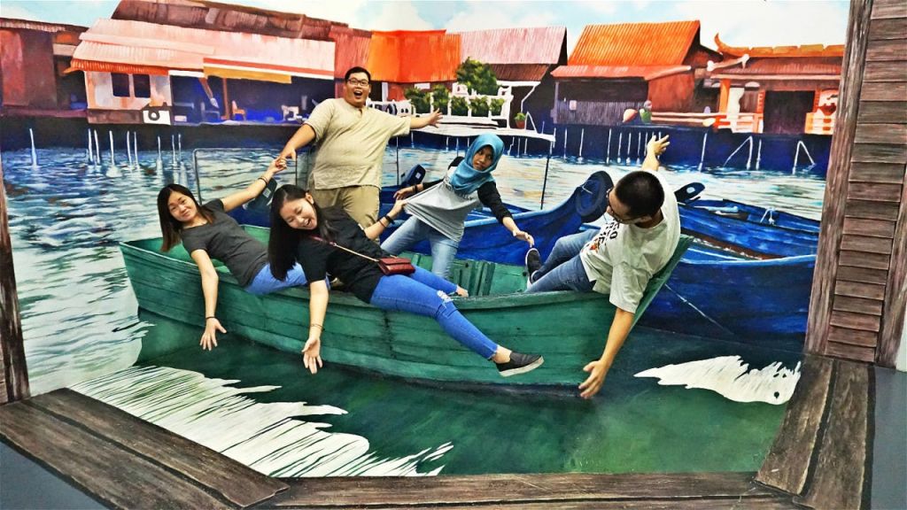 주택과 선착장이 있는 수변 풍경의 벽화에 맞서 녹색 보트를 타고 장난스럽게 포즈를 취하는 네 사람.