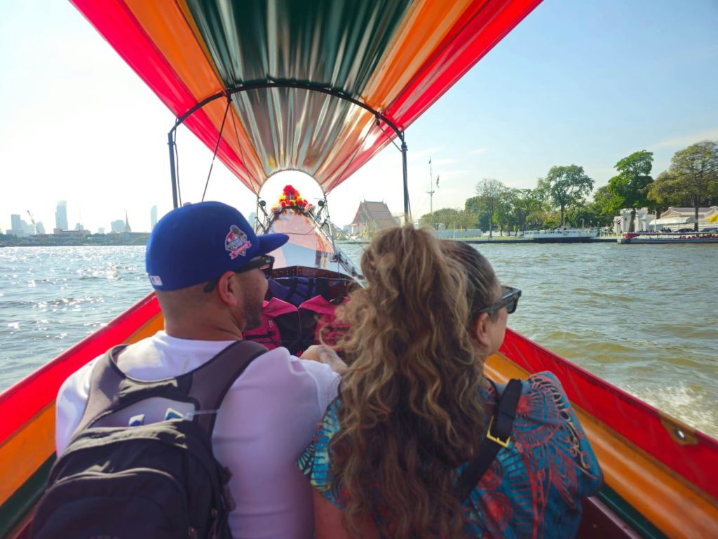 두 사람은 형형색색의 캐노피가 깔린 보트에 앉아 나무와 건물을 배경으로 맑은 하늘 아래 강을 거닐고 있습니다.