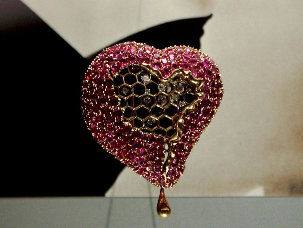 분홍색 보석으로 장식된 하트 모양의 예술 작품으로, 6각형 메쉬 디자인과 하단의 눈물방울 모양의 금색 요소가 특징입니다.