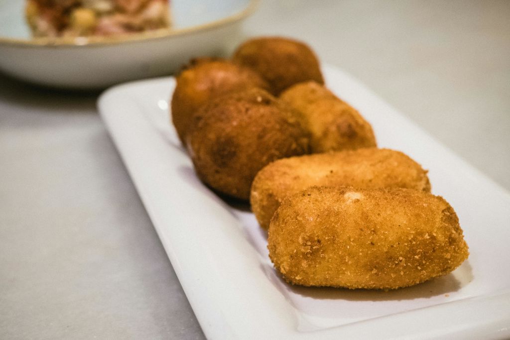 조리대에 여섯 개의 황금빛 갈색 크로켓이 담긴 흰색 접시입니다. 배경에는 그릇이 부분적으로 보입니다.