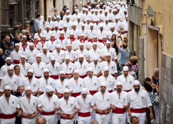 좁고 붐비는 거리를 따라 걷는 붉은 띠와 머리 덮개가 있는 흰색 의상을 입은 많은 사람들이 있습니다.