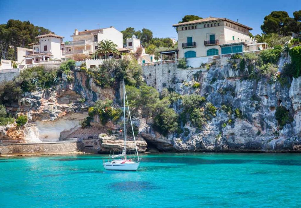 집과 녹지가 우거진 바위 절벽 근처의 맑은 청록색 물 위에 하얀 범선이 떠 있습니다.